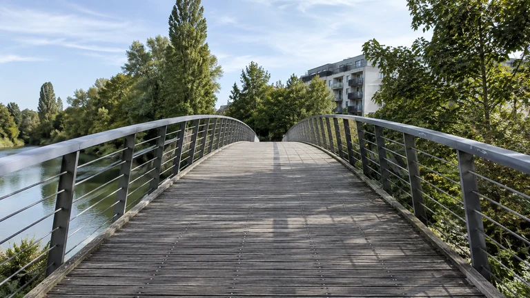 Une passerelle piétonne traverse un cours d’eau entre les arbres et les immeubles. Illustration générée par IA pour rbreak.com.